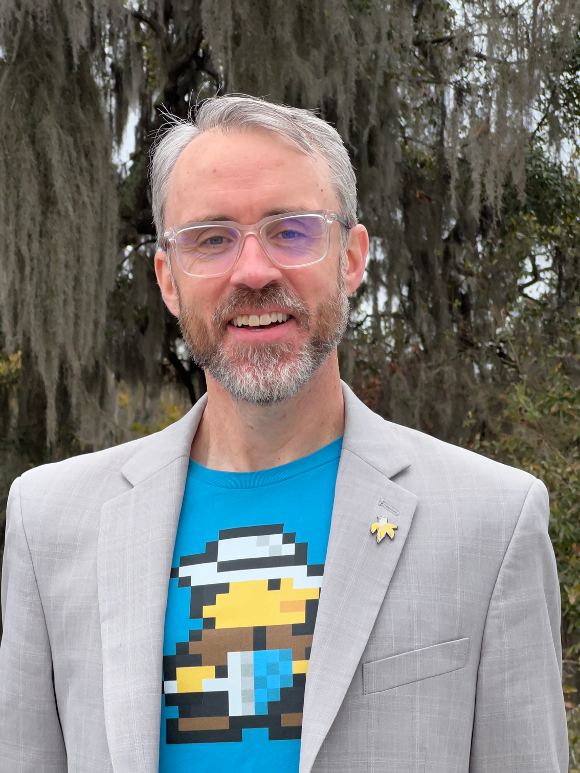 Brian Canada outdoors, wearing glasses, a light gray blazer, and a bright blue graphic shirt.
